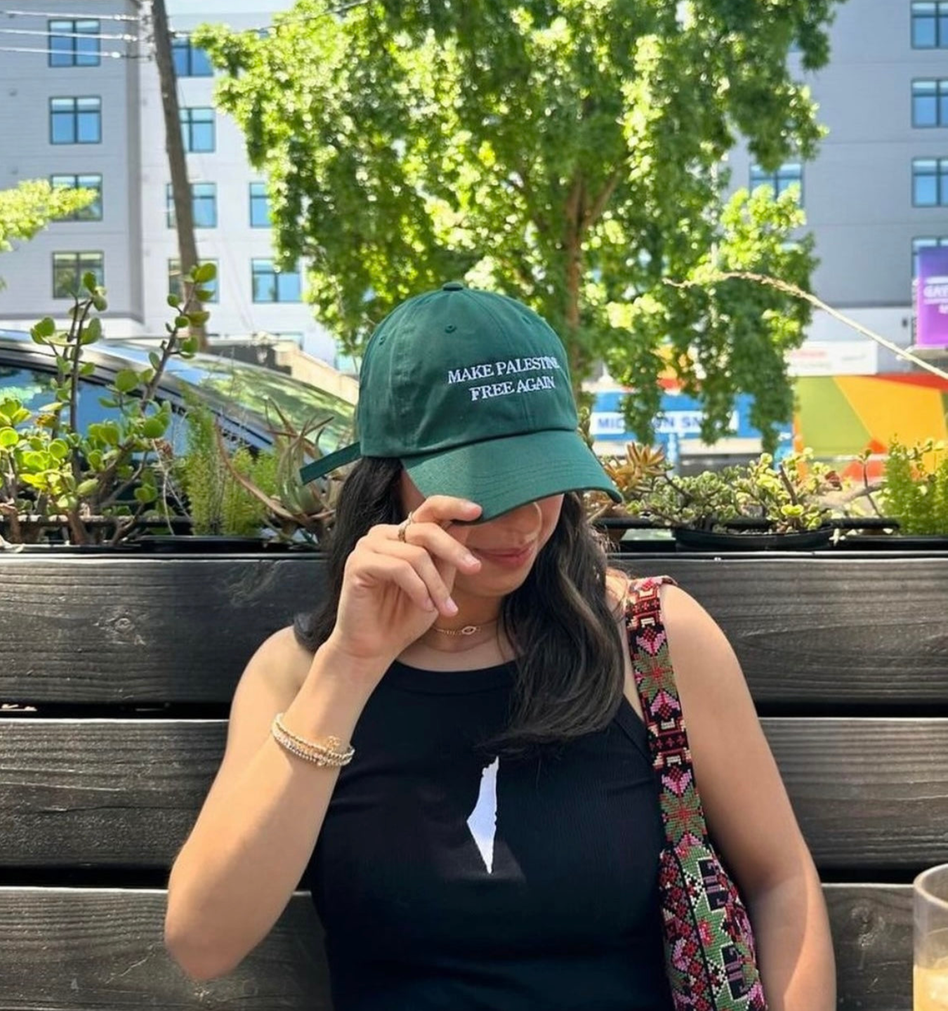 Person wearing a green cap with text, sitting outdoors with plants and buildings in the background